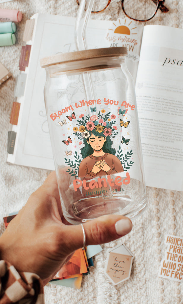Image of a woman holding a can cup glass with Bloom Where You Are Planted design, featuring a young woman surrounded by flowers and butterflies. In the background, there is an open Bible with highlighters, stickers, and reading glasses, symbolizing Bible study and reflection.