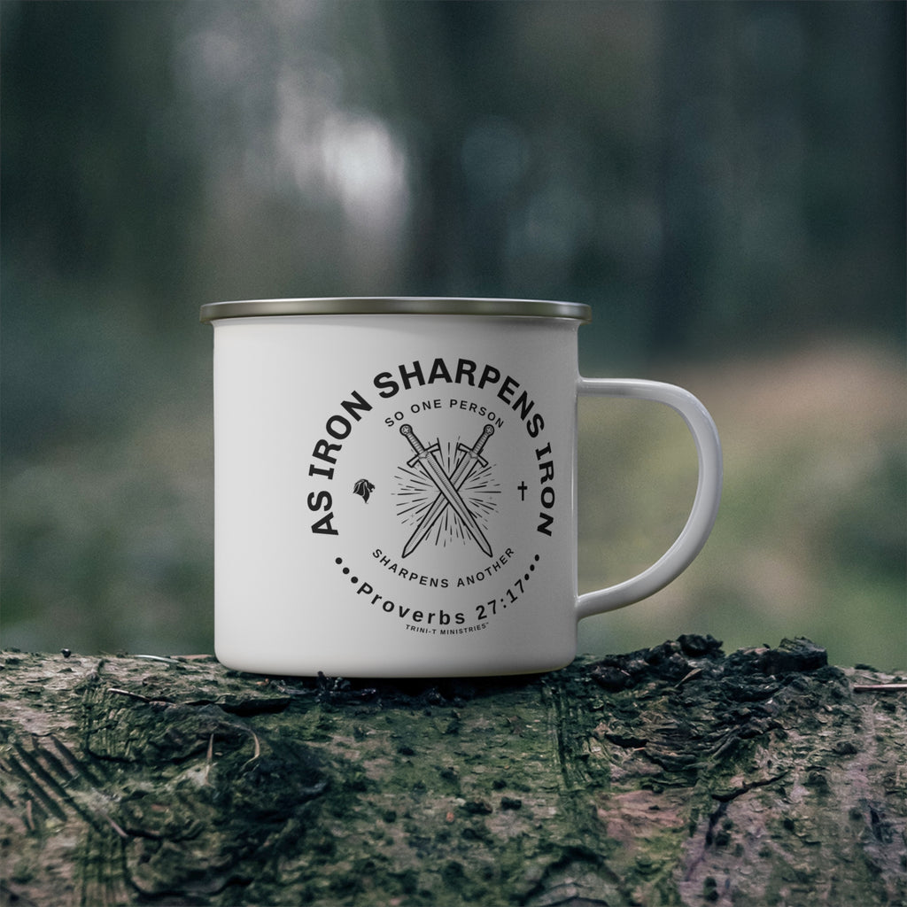 White enamel camping mug with Iron Sharpens Iron logo, resting on a mossy log in an outdoor, natural setting.