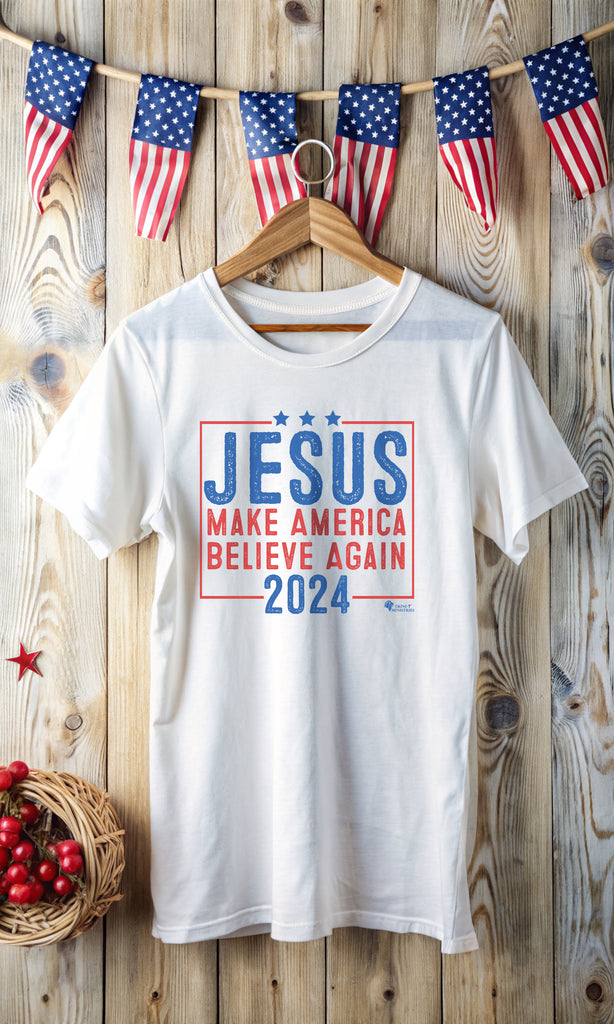 A White T-shirt with Trini-T Ministries' Jesus 2024 - Make America Believe Again design, hanging in front of a wall with American flag decorations. Embrace your faith and share a powerful message with our "Jesus 2024 - Make America Believe Again" unisex t-shirt. Designed for Christians who put Jesus above politics and candidates, recognizing Him as our true Savior. This tee blends a playful spoof of political campaigns with a unifying call to believe in Him.
