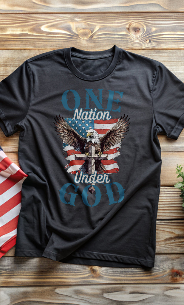 Trini-T Ministries' One Nation Under God - Eagle - Black T-shirt on wood with an American flag to the side. Wear your faith and patriotism boldly with our "One Nation Under God" patriotic T-shirt. It features a striking design of an American Bald Eagle, a Cross, and the iconic American flag.