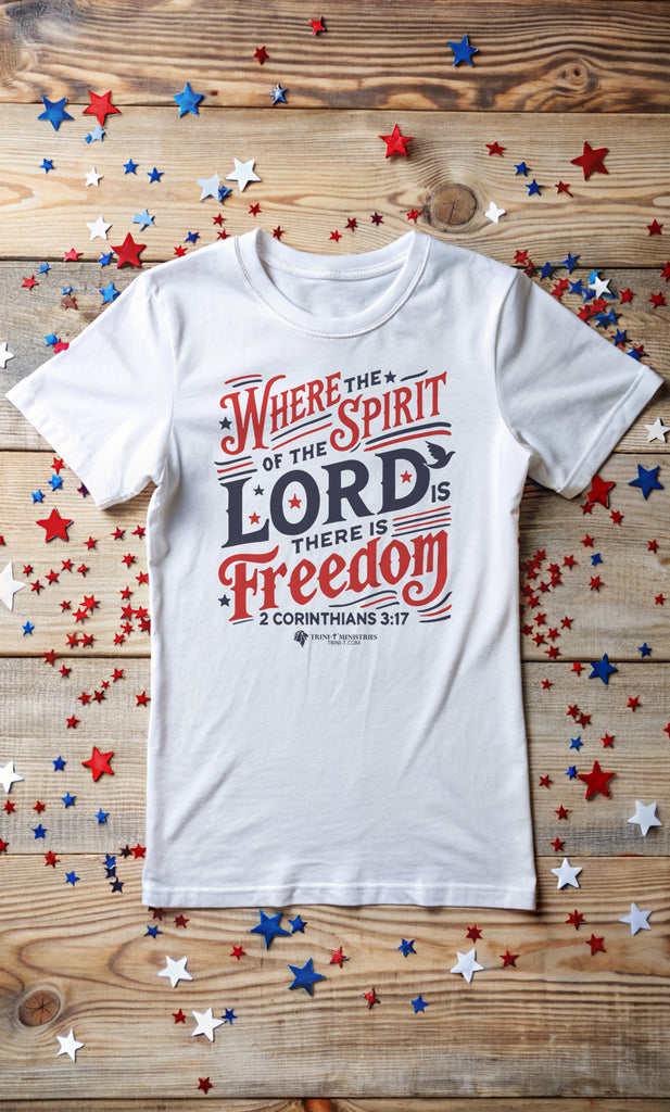 White t-shirt with 'Where the Spirit of the Lord is, there is freedom - 2 Corinthians 3:17' design, laying flat on a wooden table surrounded by red, white, and blue star-shaped confetti. Celebrate true freedom with this Christian, Scripture-inspired patriotic tee.