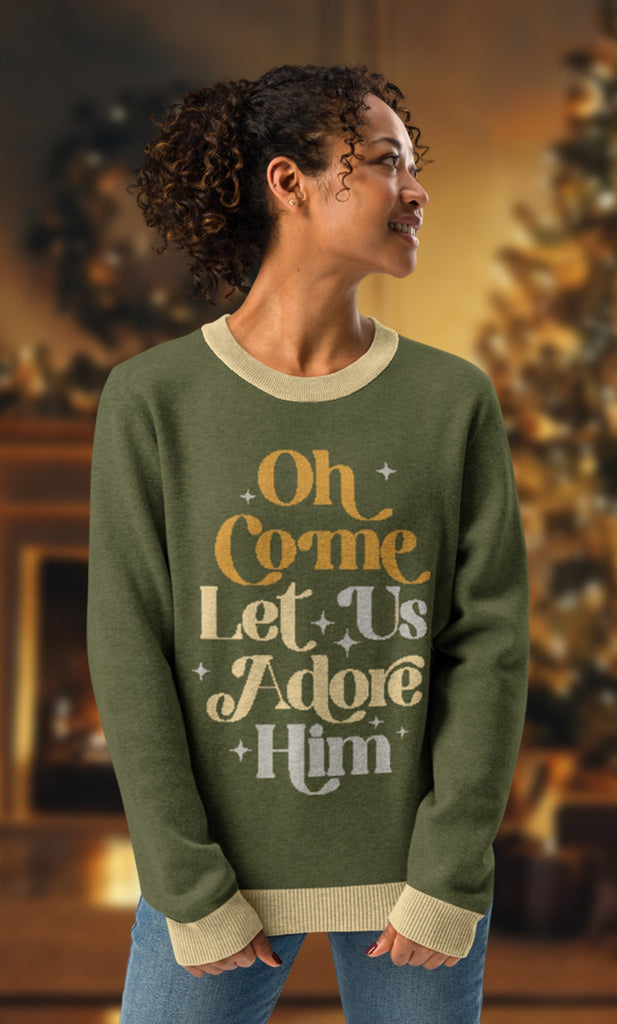 Model wearing 'Oh Come Let Us Adore Him' knitted Christmas sweater in olive green with beige trim, featuring retro-style text design in gold and cream. Christian-themed holiday apparel, unisex, cozy fit for winter and festive gatherings. Background includes a warmly lit room with a Christmas tree, evoking a cozy holiday atmosphere.