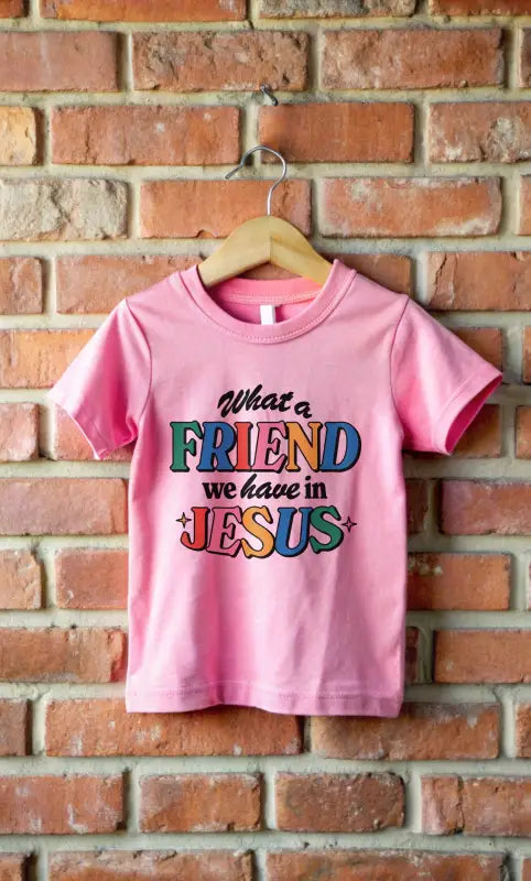 A pink Friend in Jesus - Toddler T-shirt hanging on a brick wall with vibrant, multi-colored text reading 'What a Friend we have in Jesus.' This Christian-themed shirt is perfect for little ones to boldly share their faith. Made from soft, breathable fabric, it’s ideal for daily wear or church. The John 15:15-inspired design serves as a sweet reminder of Jesus' friendship.