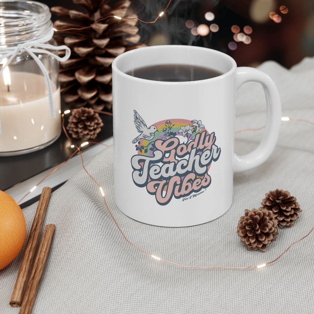 A white ceramic mug with the Godly Teacher Vibes design from Trini-T Ministries on a table with a candle, lights, and pine cones, suggesting a Christmas setting.