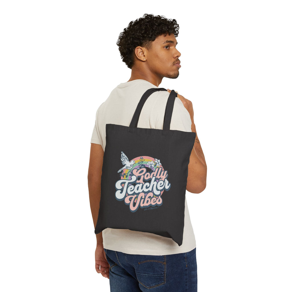 A male model carrying a black Godly Teacher Vibes Tote bag from Trini-T Ministries over his shoulder.