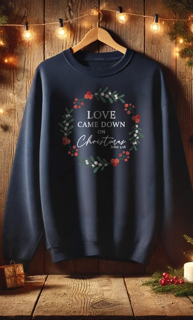 Love Came Down on Christmas navy blue sweatshirt hanging in a cozy rustic setting with warm lights, showcasing John 3:16 inspired wreath design.