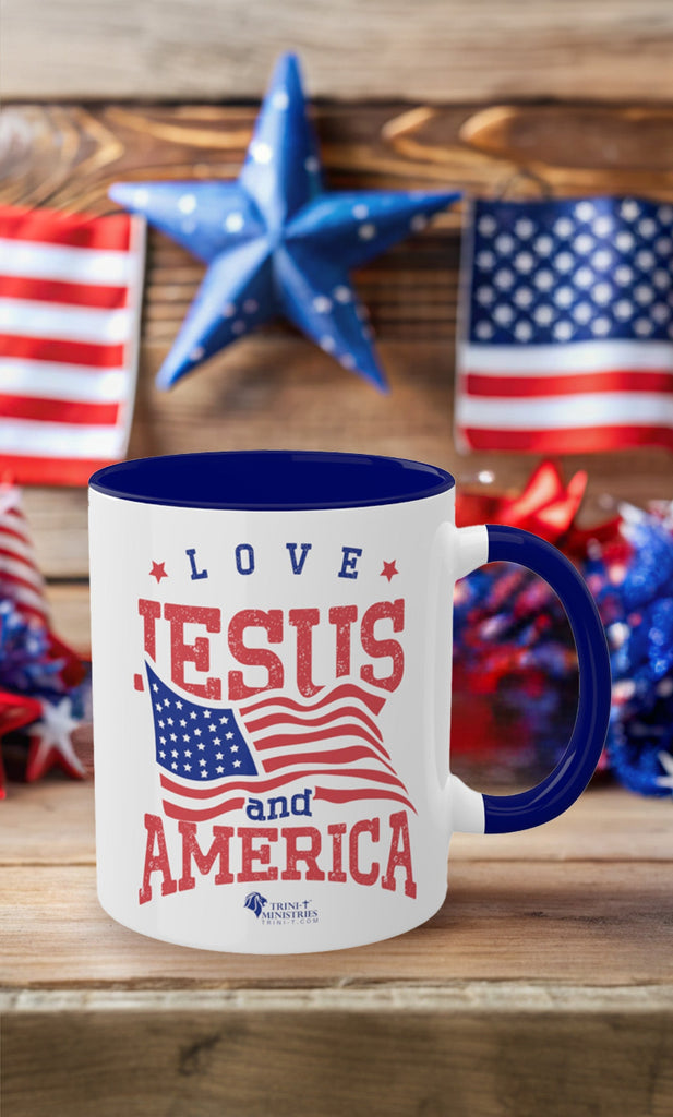 Trini-T Ministries' Love Jesus and America accented handle 11oz Blue mug on a table with American patriotic decorations in the background. Celebrate your faith and patriotism every morning with our "Love Jesus and America" 11oz Mug. This Christian Patriotic Ceramic Coffee Cup features a bold American flag design and the heartfelt message "Love Jesus and America," perfect for showing your love for God and country.