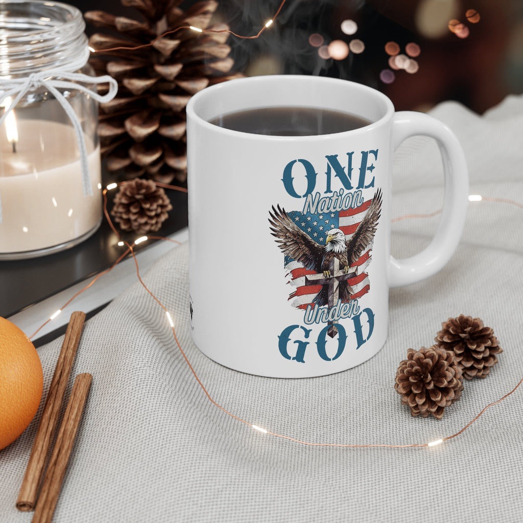 Contextual view of Trini-T Ministries' One Nation Under God - Eagle - Mug on a table with pine cones, a candle, lights, cinnamon, and an orange, filled with a hot drink. Enjoy your favorite hot beverage with a touch of faith and patriotism using our "One Nation Under God" patriotic ceramic mug. This classic white mug is perfect for coffee, tea, and hot chocolate enthusiasts. It features a striking design with an American Bald Eagle, a Cross, and the iconic American flag.